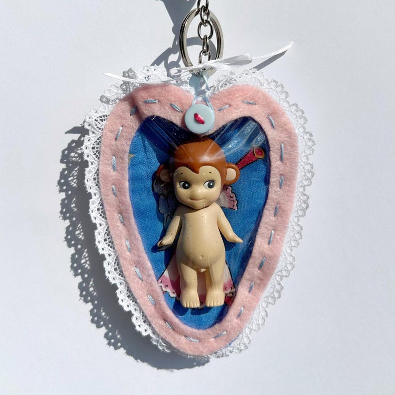 May include: A handmade keychain with a small, nude monkey figurine inside a pink felt heart. The heart is trimmed with white lace and a blue button. A silver chain and ring are attached for hanging.