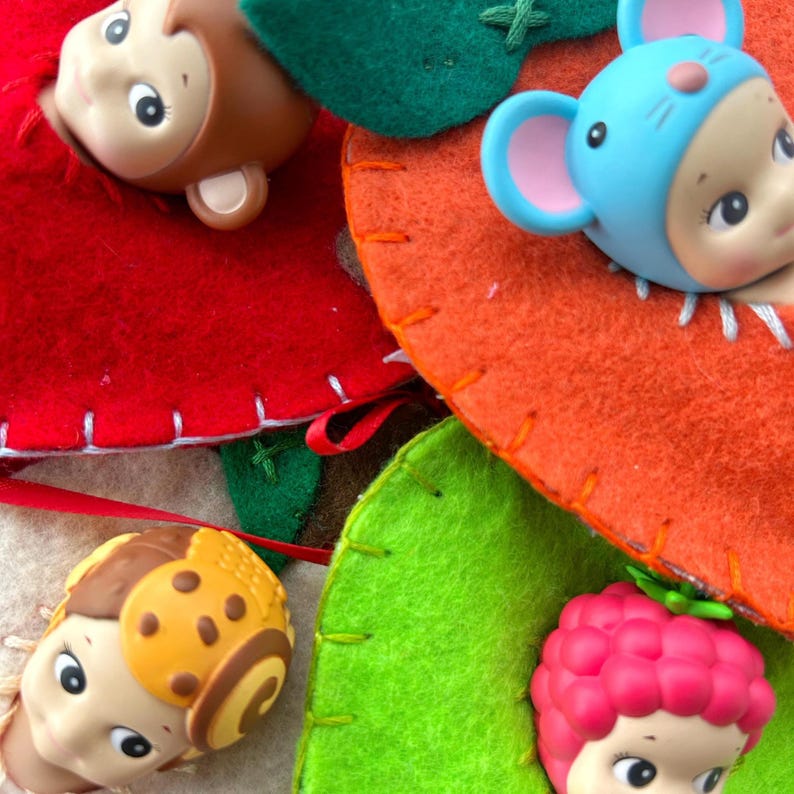May include: A colourful felt toy with four sections in red, orange, green, and beige. Each section has a doll head attached, including a monkey, a mouse, a ladybug, and a berry-themed doll. The felt is stitched together with white thread.