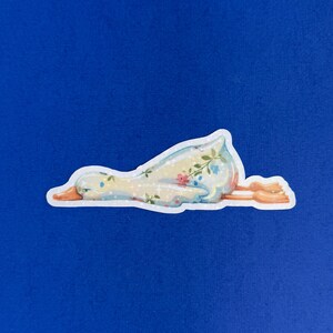 May include: A die-cut sticker of a goose lying flat on its stomach. The goose has a light blue and white body decorated with a delicate floral pattern of small pink and blue flowers, green leaves, and tiny white sparkles. It has orange feet.