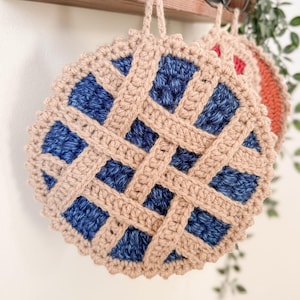 May include: Crocheted round pot holders in a pie design. The pot holders have a tan lattice design over a blue center. A second pot holder is visible with a red center. They hang from a wooden shelf with a plant.