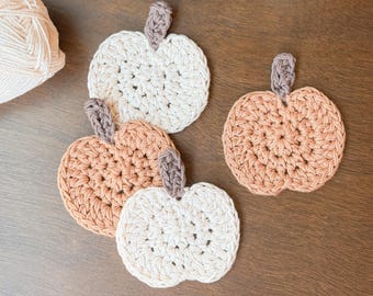 May include: Four handmade crochet pumpkin coasters in shades of cream and peach, each with a brown stem. The coasters are arranged on a wooden surface, suggesting a fall theme. Yarn balls are visible in the background.