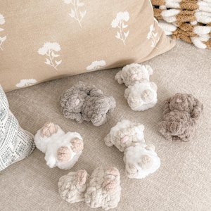 May include: A collection of small, handmade crochet lamb toys in various shades of white, gray, and brown. The lambs have fluffy bodies and are arranged on a neutral-toned surface, with a floral patterned pillow in the background.