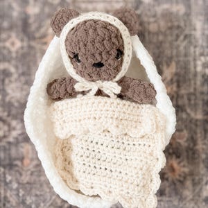 May include: A handmade crochet baby bear in a white cocoon. The bear is brown with a white bonnet tied with a bow. The cocoon is white with a scalloped edge blanket. The item is designed for a baby.