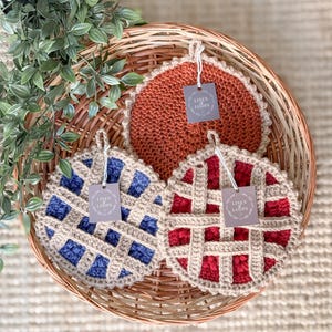 May include: Three crocheted pie-shaped pot holders in a wicker basket. One is rust-colored, another is blue, and the third is red. Each has a lattice design and a tag that reads "LINEN LOOPS". The pot holders are handmade.