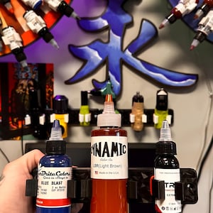 May include: Three bottles of tattoo ink, two blue and one orange, are displayed on a black ink holder. The bottles are labeled "Brite Colors Goes in Stays in Blue Blast", "Dynamic Color Co. LBR(Light Brown)", and "Advanced Extra".