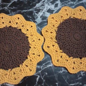 May include: Two handmade crochet sunflower coasters. Each coaster features a dark brown center and a yellow scalloped edge, resembling a sunflower. These decorative coasters are perfect for adding a touch of nature to your home decor.