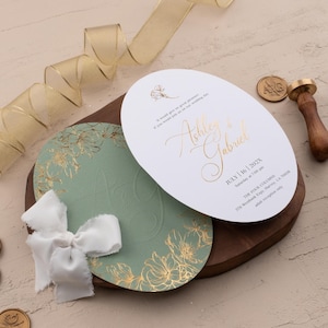 Könnte beinhalten: Elegante Hochzeitseinladungen mit goldenen Folienakzenten. Die Einladungen zeigen die Namen "Ashley & Gabriel" und das Datum "16. Juli 202X". Ein weißes Band und goldene Wachssiegel ergänzen das elegante Design. Ein goldenes Band ist ebenfalls vorhanden.