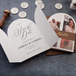 Op de afbeelding: Witte trouwuitnodiging met de tekst "Save the Date" in elegant schrift. De uitnodiging bevat de namen Ashley & Gabriel, de trouwdatum en locatie. Een bruine envelop en foto's zijn zichtbaar.