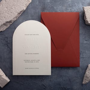 Pode incluir: Um convite de casamento branco sujo com um topo arqueado e texto em relevo, incluindo os nomes Ashley e Gabriel e a data de 6 de agosto de 2023. Um envelope vermelho escuro está ao lado do convite. O convite está em uma superfície cinza escuro com detalhes em pedra.