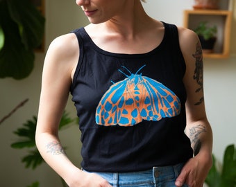 May include: A woman wearing a black tank top with a blue and orange moth graphic. She is wearing blue denim jeans and has short blonde hair.