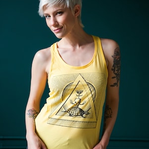 Könnte beinhalten: Eine Frau mit kurzen, platinblonden Haaren trägt ein gelbes Tanktop mit einem schwarz-weißen Grafikdruck eines Wesens in einem Dreieck. Sie trägt auch schwarze Jeans-Shorts.