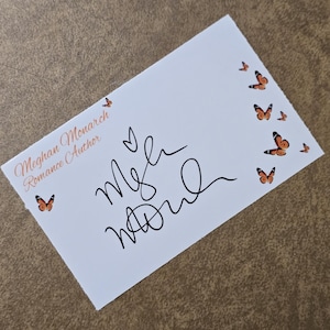 May include: A white card with the text "Meghan Monarch Romance Author" and a handwritten signature. The card is decorated with small orange butterflies. A heart symbol is drawn above the signature. The card is on a brown surface.