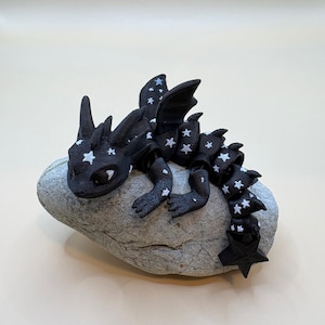 Könnte beinhalten: Eine schwarze Drachenfigur mit weißen Sternmustern auf Flügeln, Rücken und Schwanz, die auf einem grauen Stein ruht. Der Drache hat detaillierte Merkmale und steht vor einem neutralen Hintergrund.