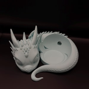 Könnte beinhalten: Ein hellblauer Keramik-Drachen-Pflanztopf mit einem eingerollten Schwanz und einem ausgehöhlten Körper zum Pflanzen. Der Drache hat ein detailliertes Gesicht mit geschlossenen Augen und einen strukturierten Körper.