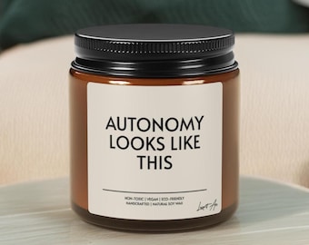 May include: A brown glass jar with a black lid, displaying a white label with the words "AUTONOMY LOOKS LIKE THIS." The label also states the candle is non-toxic, vegan, eco-friendly, handcrafted, and made with natural soy wax. The jar is on a light-coloured surface.