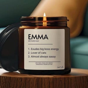 Puede incluir: Un tarro de vela de vidrio marrón con una etiqueta blanca que dice "EMMA [eh-muh] sustantivo 1. Desprende energía de jefe 2. Amante de los gatos 3. Casi siempre descarada" y el texto "NO TÓXICO | VEGANO | ECOLÓGICO HECHO A MANO | CERA DE SOJA NATURAL".