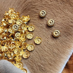 May include: A pile of gold-coloured metal studs with a decorative design. The studs are scattered on a brown furry surface.