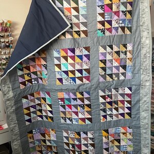 May include: A grey quilt with a patchwork design made of colourful triangles. The quilt is made of many different fabrics and colours, including blue, purple, pink, yellow, and green. The quilt is folded in half and is being held by a person's hand.