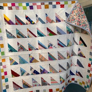 May include: A patchwork quilt featuring a white background with rows of multi-coloured fabric triangles. The border consists of a grid of small, vibrant squares in various colours. The reverse side displays a floral pattern with pink and green.