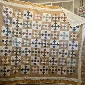 May include: A beige quilt with a patchwork design featuring a variety of floral and geometric patterns. The quilt is made up of squares with a diamond pattern in each square. The quilt is folded and displayed on a white surface.