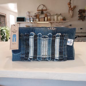 May include: A denim pouch with a patchwork design of various shades of blue denim. The pouch has a tag that reads "The Rancher Pants Bag Co." and a small white tag with a blue flower. The pouch is sitting on a white surface.