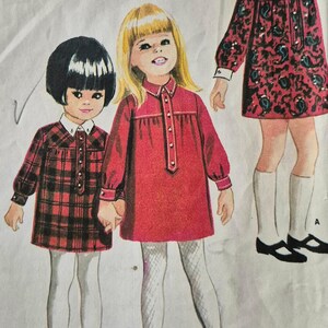 Può includere: Un'illustrazione di cartamodello per un vestito da bambino in tre versioni. I modelli presentano maniche lunghe, un'abbottonatura frontale e un colletto bianco. Una versione è rosso tinta unita, una a quadri rossi e neri e una floreale.