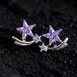 Peut inclure: Boucles d'oreilles argentées en forme d'étoiles avec des accents de pierres précieuses violettes. Les boucles d'oreilles présentent un design incurvé avec trois étoiles de tailles différentes. L'image est sur un fond sombre et texturé. Le texte "Rivenpunk Artisans" est visible.