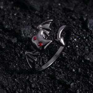 Puede incluir: Un anillo de metal negro con forma de murciélago y dos pequeñas piedras circulares rojas como ojos. El anillo presenta un diseño de banda abierta con un extremo en punta. El texto RivenPunkArtisans está impreso en blanco en la parte inferior.