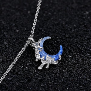May include: A silver necklace with a crescent moon pendant. The moon pendant is blue with white accents and is surrounded by a silver decorative frame. The necklace is on a dark, textured surface. The text "RivenPunkArtisans" is at the bottom.
