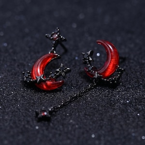 May include: A pair of celestial-themed earrings featuring red crescent moons and black star accents. The earrings have a dark, gothic aesthetic, with a dangling chain on one earring. The brand name "RivenPunkArtisans" is visible at the bottom.