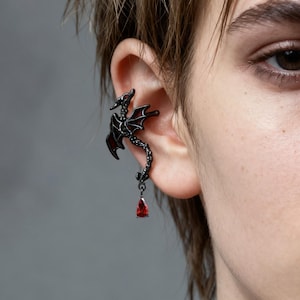 May include: Black dragon ear cuff with a red gemstone pendant. The dragon has detailed wings and a long, winding body. The ear cuff is worn on the ear, with the dragon wrapping around the ear's edge.
