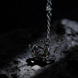 May include: A silver necklace pendant with a celestial design. The pendant features a crescent moon, stars, and a planet-like ring, with iridescent details. The chain is fine, and the background is dark.
