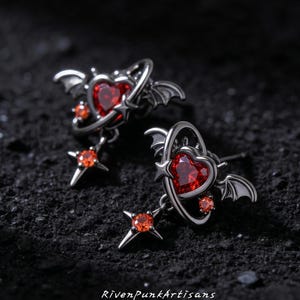 May include: A pair of silver earrings with a gothic design. Each earring has a heart-shaped red gemstone, encircled by a halo with bat wings and a star. A smaller red gem and a star-shaped charm hang below. The text "RivenPunkArtisans" is at the bottom.