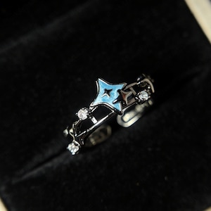 May include: A silver-toned ring with a blue star design and small clear gemstones. The ring features an open design with a central blue star and is set against a dark background. The ring is an adjustable size.