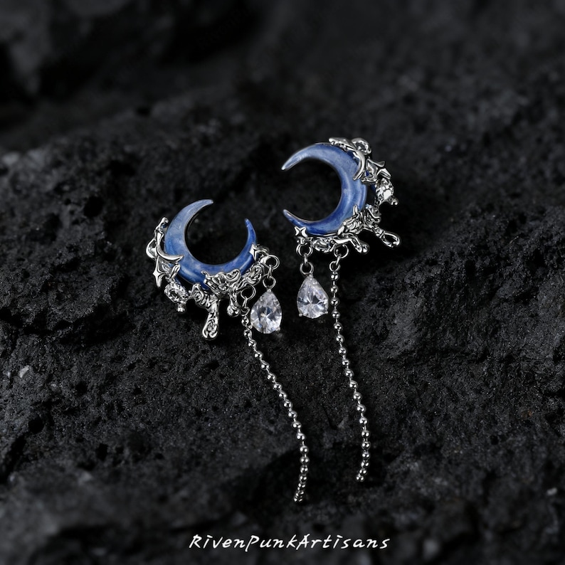 May include: A pair of silver-toned earrings featuring a crescent moon design with a blue enamel finish. Each earring has a dangling teardrop-shaped crystal and a delicate chain. The earrings are set against a dark, textured background.