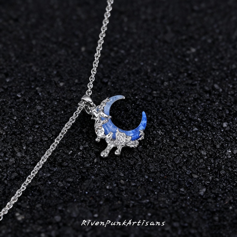 May include: A silver necklace with a crescent moon pendant. The moon pendant is blue with white accents and is surrounded by a silver decorative frame. The necklace is on a dark, textured surface. The text "RivenPunkArtisans" is at the bottom.