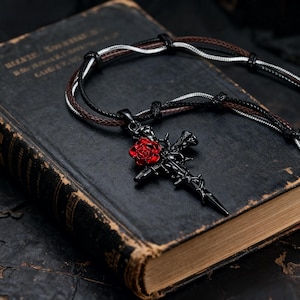 Op de afbeelding: Zwarte kruisvormige hangerketting met een rode roos. De ketting heeft een koord met meerdere strengen. De hanger ligt op een donker, antiek boek.