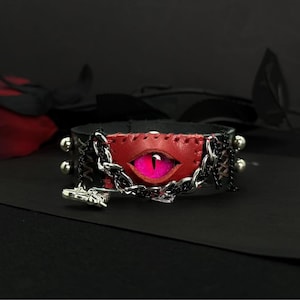 Pode incluir: Gargantilha de couro preta com um centro de couro vermelho com um design de olho rosa. A gargantilha é adornada com tachas prateadas, uma corrente prateada e um pequeno pingente. O design tem um tema gótico ou de fantasia.