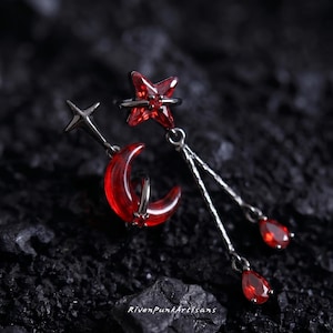 May include: A pair of asymmetrical earrings featuring celestial designs. One earring has a silver star and a red crescent moon. The other earring has a red star and two dangling red teardrops. The earrings are set against a dark, textured background.