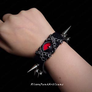 May include: Black leather bracelet featuring a red eye design with silver chain accents and metal spikes. The bracelet is a gothic accessory with a punk rock aesthetic. The text "RivenPunkArtisans" is visible at the bottom of the image.