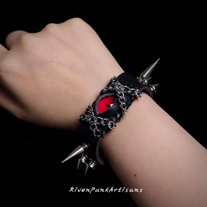 May include: Black leather bracelet featuring a red eye design with silver chain accents and metal spikes. The bracelet is a gothic accessory with a punk rock aesthetic. The text "RivenPunkArtisans" is visible at the bottom of the image.