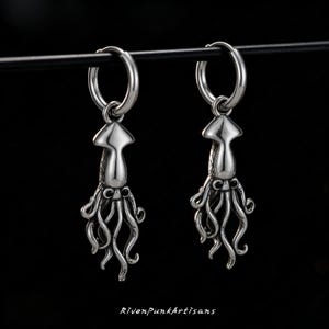 May include: A pair of silver-colored hoop earrings, each featuring a detailed squid pendant. The pendants have a stylized design with tentacles and a rounded head. The earrings hang against a black background. The text "RivenPunkArtisans" is at the bottom.