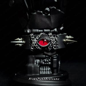May include: Black leather choker with a red eye and silver chain and spike accents. The choker features the text "RivenPunkArtisans" at the bottom. The choker is a gothic accessory.