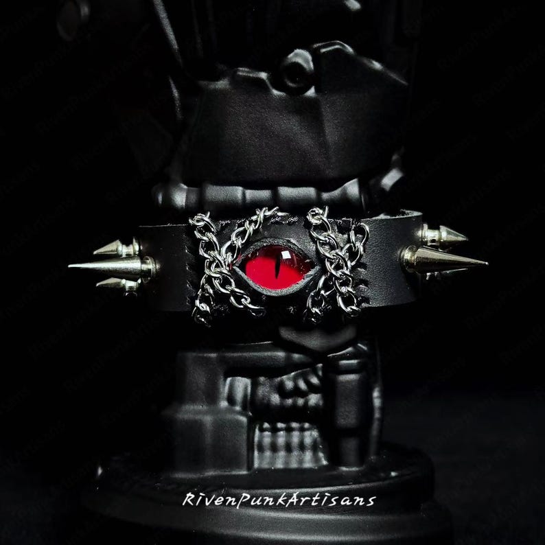 May include: Black leather choker with a red eye and silver chain and spike accents. The choker features the text "RivenPunkArtisans" at the bottom. The choker is a gothic accessory.