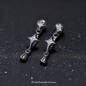 May include: A pair of silver-toned earrings with a starburst design. Each earring features a star-shaped element with a central clear stone, a small teardrop pendant, and a decorative top. The earrings are displayed against a dark, glittery background.