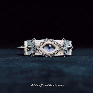 Könnte beinhalten: Handgefertigtes Lederarmband mit blauem Augendesign, silberfarbenen Kettenakzenten und Metallspitzen. Der Text "RivenPunkArtisans" ist unten zu sehen. Ein einzigartiges Accessoire.