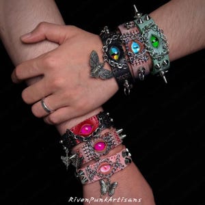 Op de afbeelding: Een verzameling leren armbanden in diverse kleuren, met oogvormige versieringen, kettingdetails en metalen spikes. Sommige armbanden hebben vlinderbedels. De armbanden worden getoond op de polsen van een persoon.
