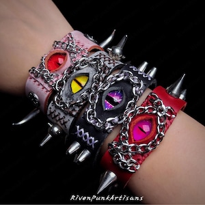 Op de afbeelding: Vier leren polsbanden in roze, grijs, zwart en rood, elk met een centraal oogontwerp, een kettingrand en metalen spikes. De ogen hebben verschillende kleuren: rood, geel, paars en roze. De tekst "RivenPunkArtisans" staat onderaan.