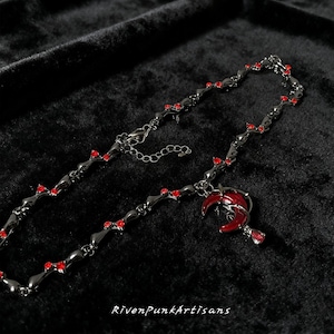 May include: A dark gray necklace with red accents and a crescent moon pendant. The necklace features small red gems and a matching red crescent moon charm. The text "RivenPunkArtisans" is visible at the bottom.