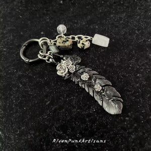 Puede incluir: Un llavero plateado con un diseño detallado de pluma adornado con pequeñas rosas. El llavero incluye un cierre, una cadena con cuentas de piedra y el texto "RivenPunkArtisans".